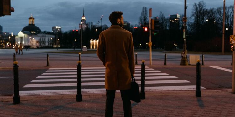 Чалавек у касцюме з невялікім чамаданам і заплечнікам стаіць на фоне гістарычнага еўрапейскага горада, разглядаючы карту.