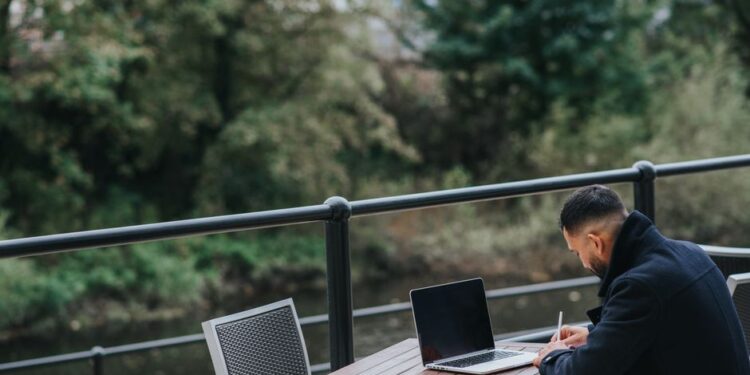 Un uomo d'affari lavora al laptop in un caffè con vista sulla città, simboleggiando la transizione tra lavoro e tempo libero.