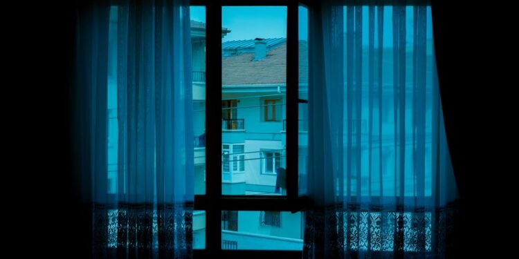 Жанчына з кубкам гарбаты сядзіць на канапе ўвечары, святло ад ноўтбука, за акном гарадскі пейзаж. Стварае адчуванне камфорту пасля працоўнага дня.