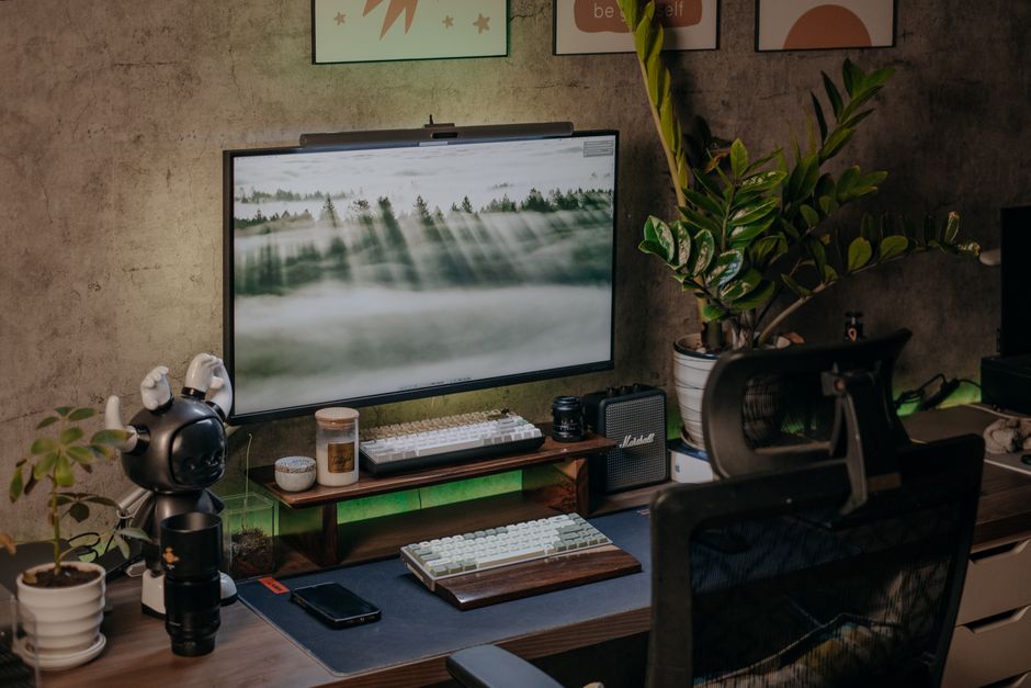 Scrivania di casa ben organizzata con monitor, tastiera, piante e luce naturale, per un ambiente di lavoro ergonomico e rilassante.