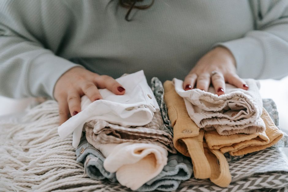 Mani di una persona che piegano con cura una maglia su un tavolo, con altri capi ben organizzati sullo sfondo.
