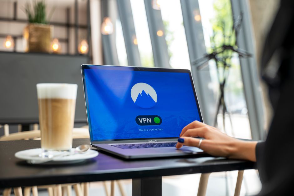 Primo piano delle mani di una persona che configura un'applicazione VPN su un laptop, seduta a un tavolo di un bar con un caffè e un telefono nelle vicinanze, suggerendo la navigazione sicura sul Wi-Fi pubblico.