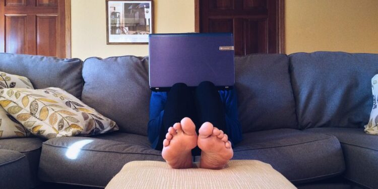 Una persona seduta sul divano che usa lo stesso laptop sia per lavorare che per rilassarsi, con una tazza di caffè e una rivista accanto.