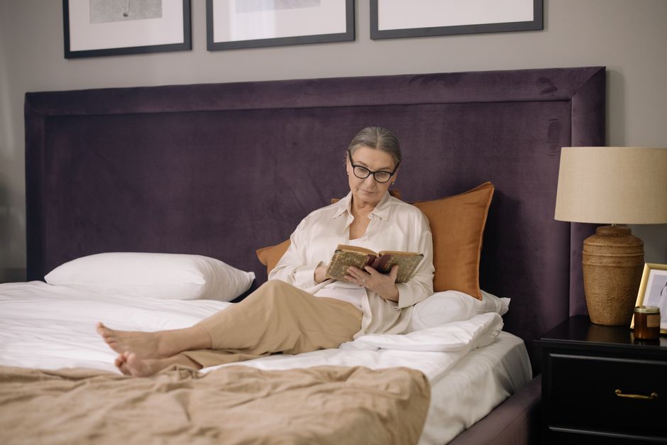 Una donna serena in un letto accogliente, legge un libro cartaceo sotto una luce soffusa, con una tisana sul comodino.