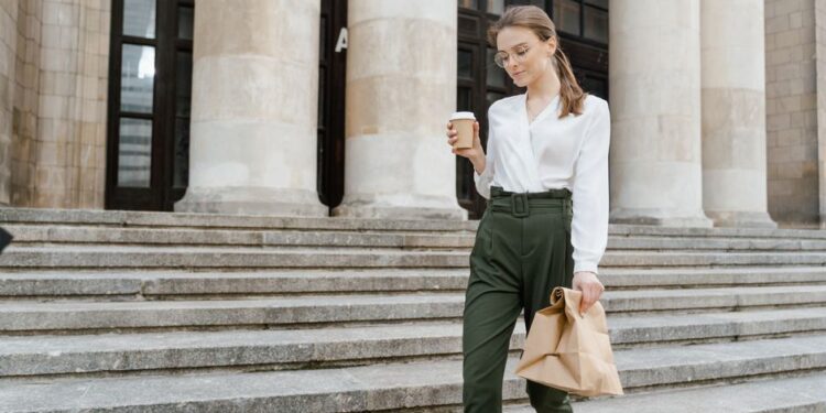 Donna con outfit smart casual elegante seduta in un caffè, sorseggiando un caffè e guardando il telefono.