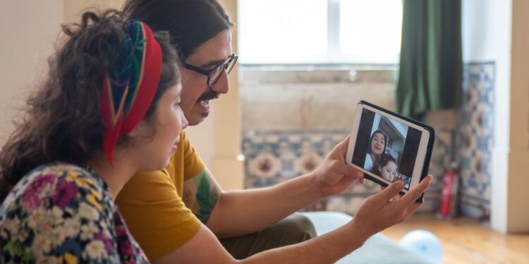 Dva přátelé se usmívají během videohovoru, jeden z nich drží tablet, zatímco druhý je vidět na obrazovce. Naznačuje přátelství na dálku.