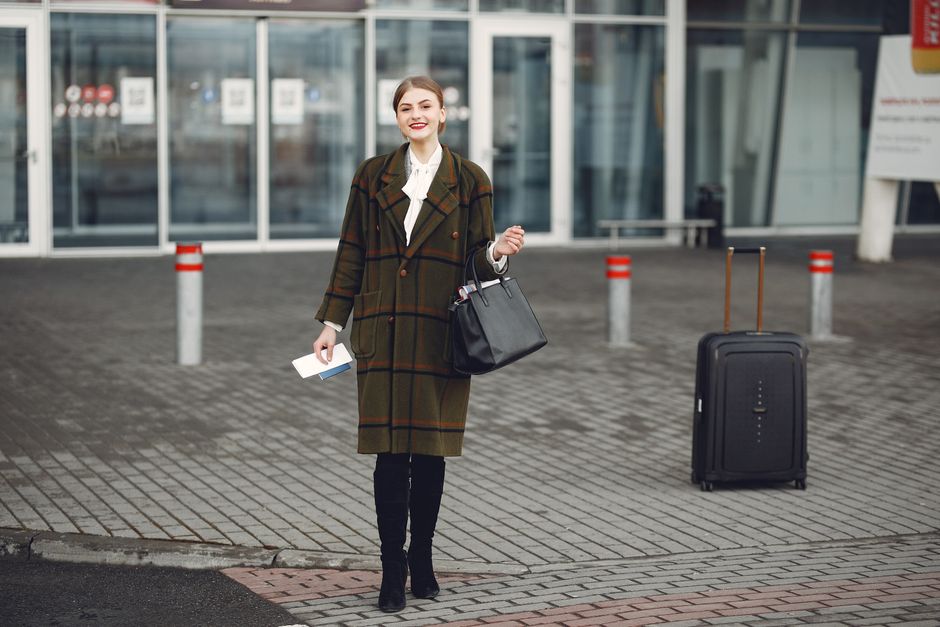 Stylová žena v elegantním kabátu a šátku s malým kufrem na letišti, připravená na cestu.