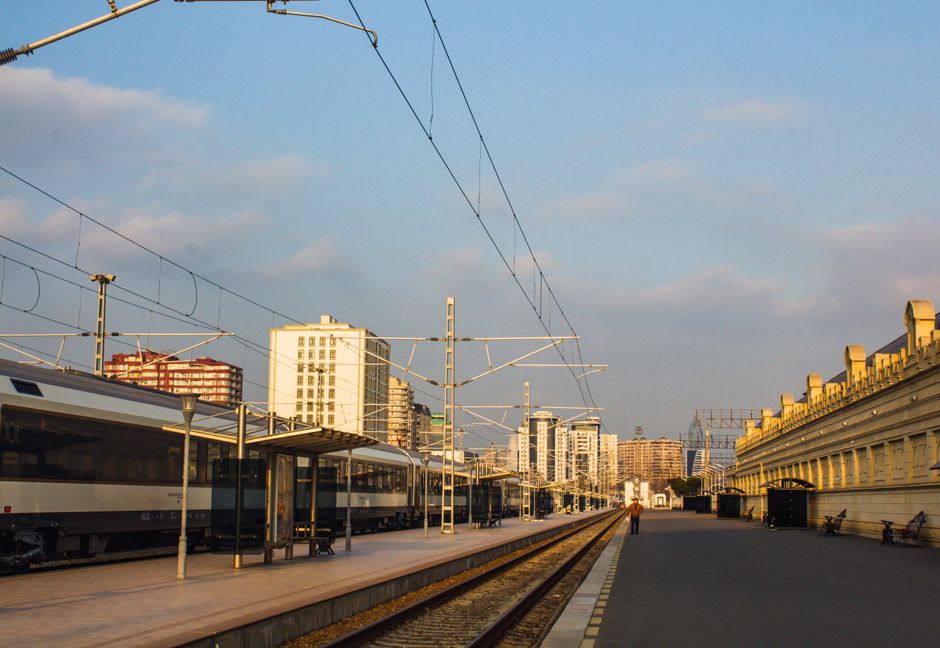 Moderní vlakové nádraží v okrajové části města s čistými perony a rychlým vlakem přijíždějícím do stanice. V pozadí se rýsují obytné budovy a zelené plochy, což ilustruje dobrou dopravní dostupnost a klidnější prostředí.
