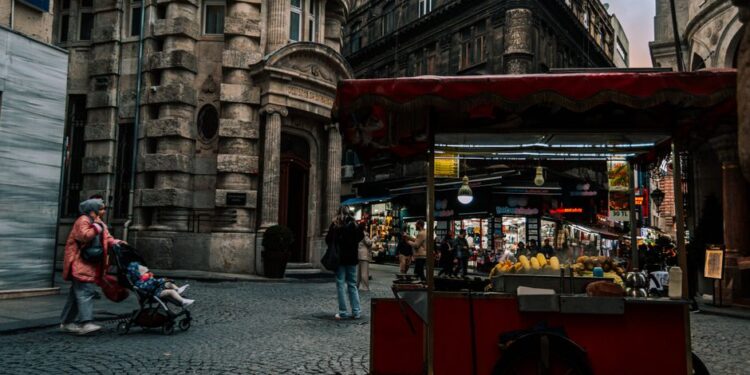Turisté s batohy a fotoaparáty procházející přeplněnou ulicí s obchody se suvenýry.